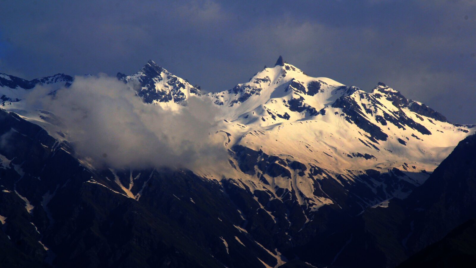 Snow-covered Shrikhand Mahadev peak above dark mountain slopes.