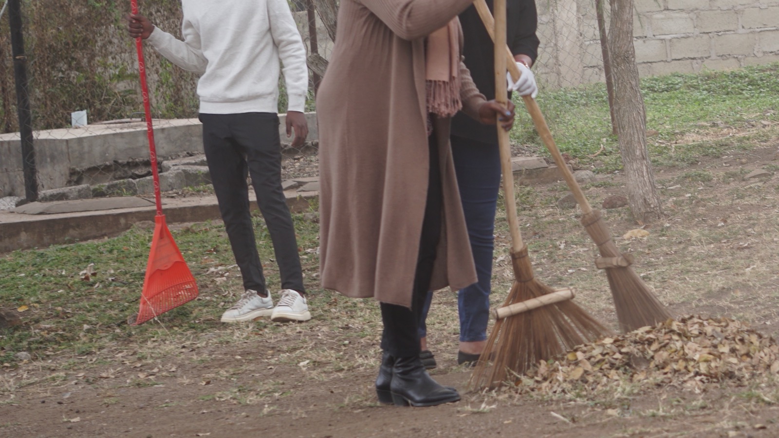 People holding brooms and a leaf rake beside a pile of dry leaves.